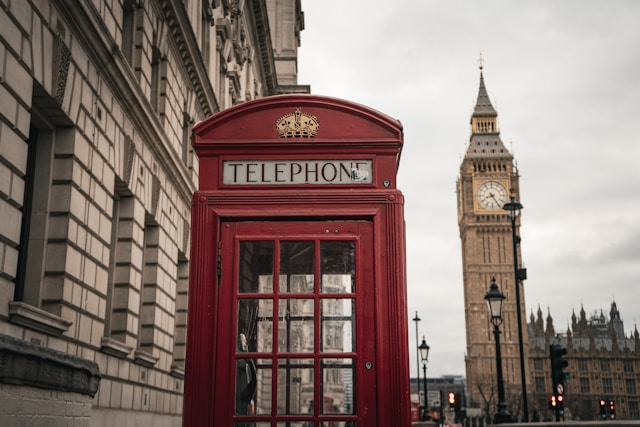 Red phone booth near Big Ben and historic building on cloudy day.