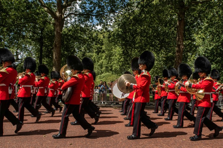Marching band in red uniforms and black hats parades through a tree-lined path.