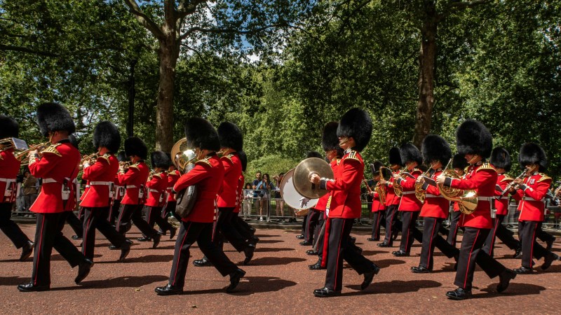 Marching band in red uniforms and black hats parades through a tree-lined path.