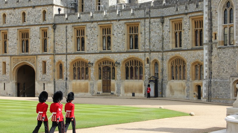 Guards in red uniforms march near a historic stone building.