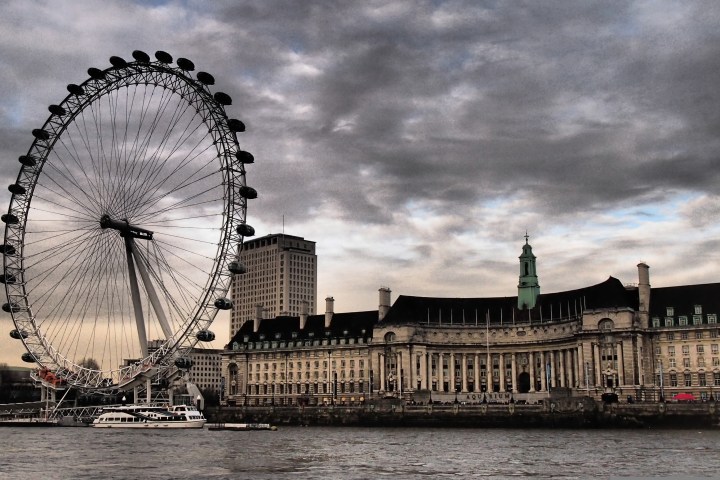 Ferris wheel near a historic building by the river under a cloudy sky.