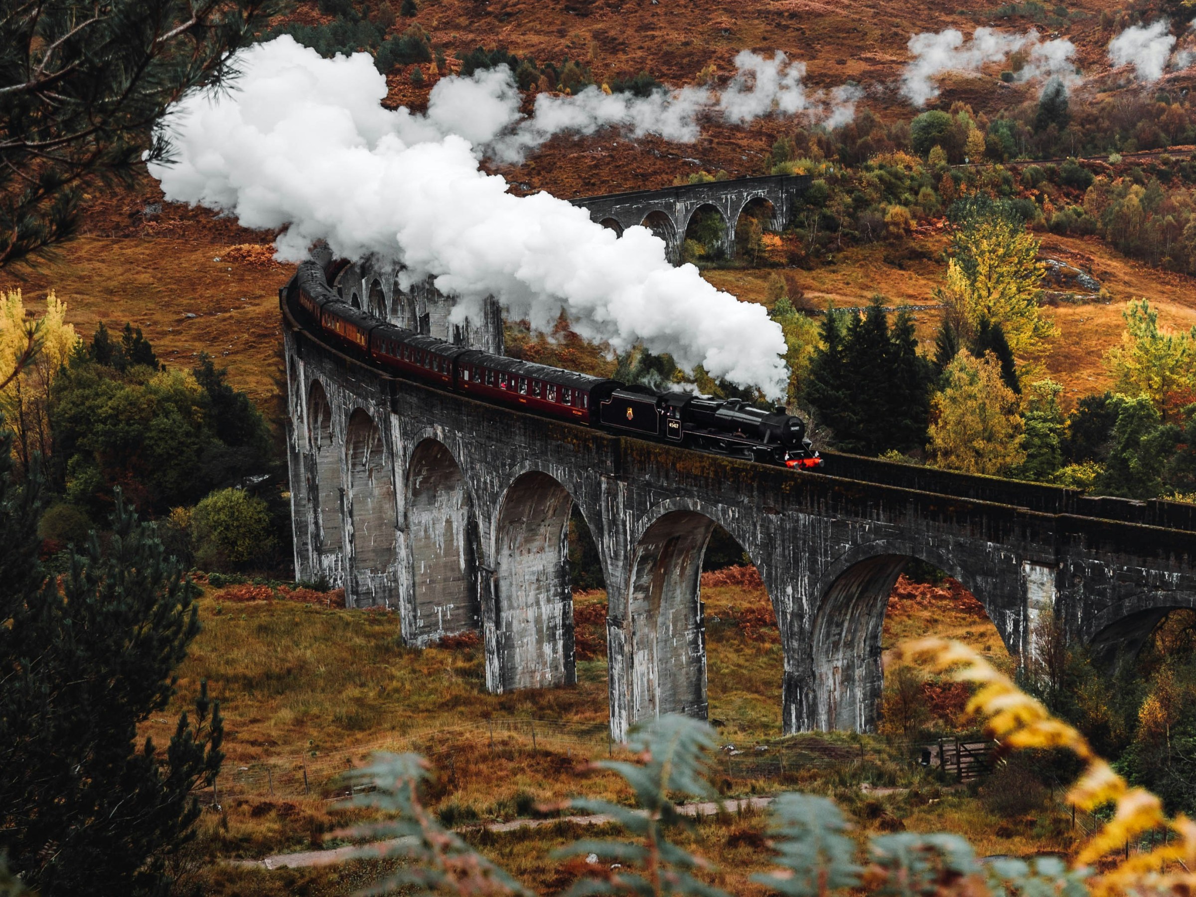 Steam train crosses a stone viaduct in a scenic, autumn landscape.