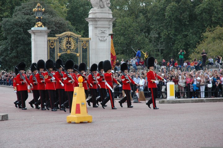 Guards in red uniforms and bearskin hats march near ornate gates as a crowd watches.
