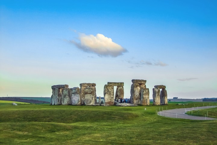 Stonehenge under a blue sky with a single cloud, surrounded by green grass.