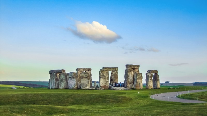Stonehenge under a blue sky with a single cloud, surrounded by green grass.