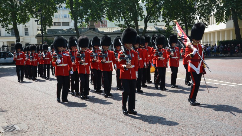 Soldiers in red uniforms and bearskin hats marching with instruments on a street.