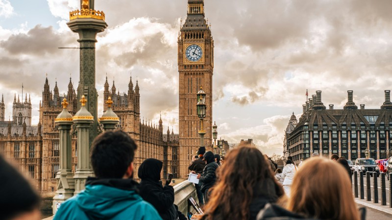 People walking on a bridge towards Big Ben under cloudy skies.