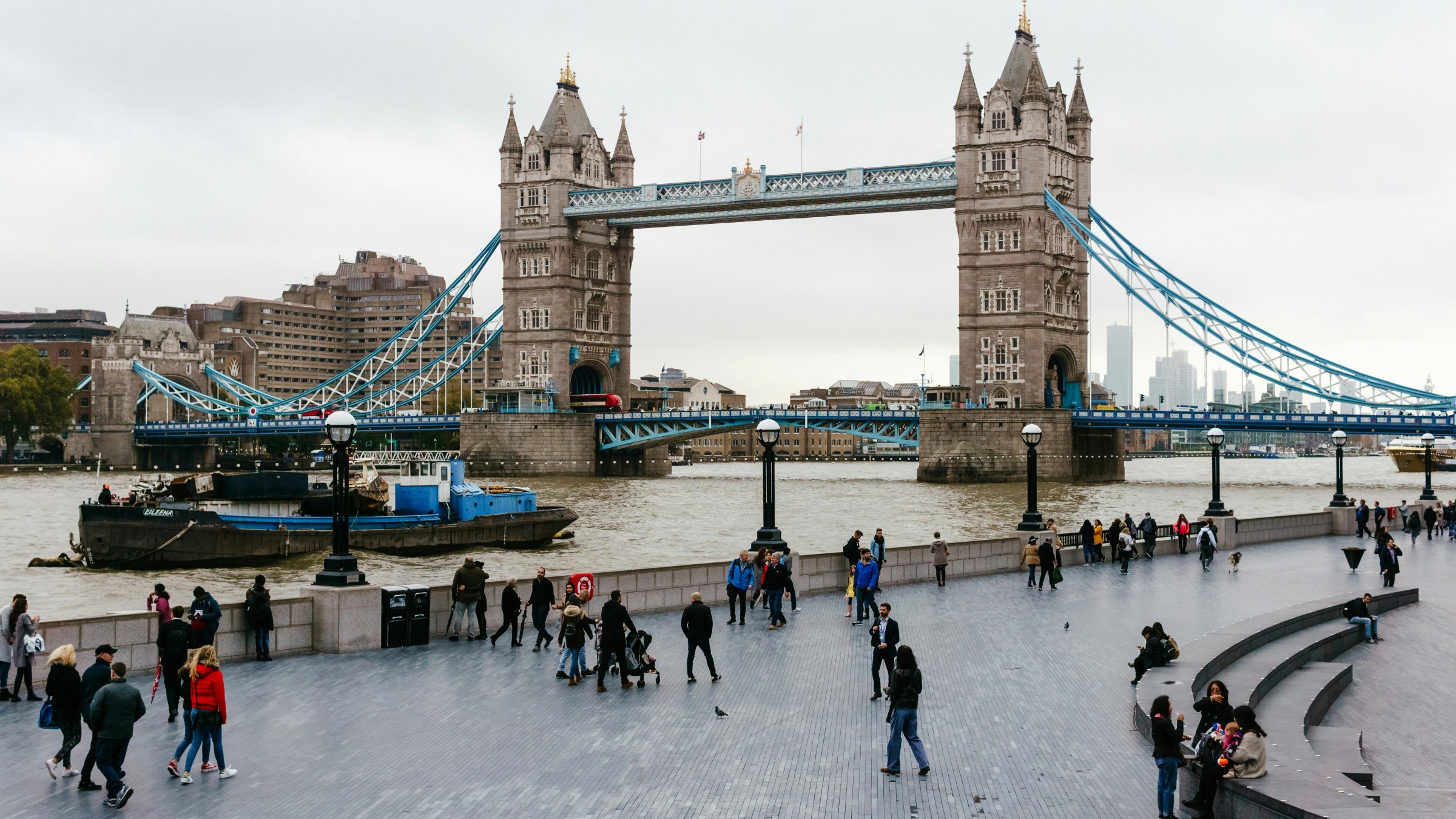 People walking near Tower Bridge over River Thames on a cloudy day.
