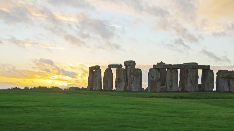 Stonehenge silhouetted against a colorful sunset sky with green grass in the foreground.