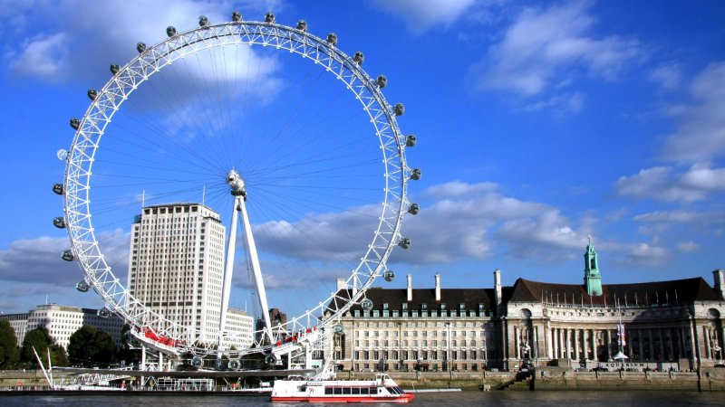 Large Ferris wheel by a river with historic buildings and a cloudy sky.