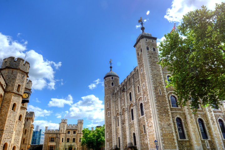 Historic stone castle towers under a clear blue sky with scattered clouds and green trees.
