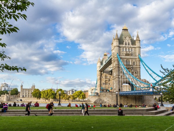 Tower Bridge in London with people sitting on grass and a partly cloudy sky.