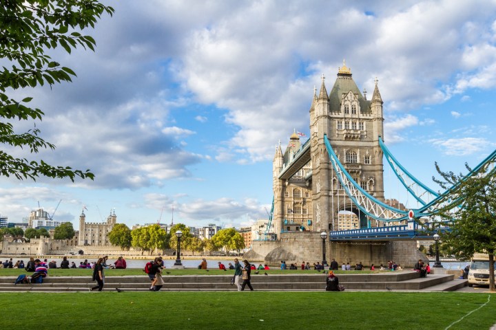 Tower Bridge in London with people sitting on grass and a partly cloudy sky.