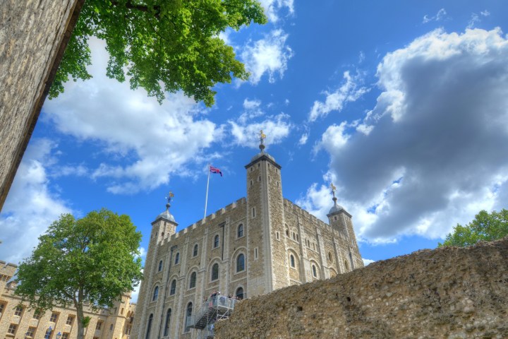Historic stone castle with flags, surrounded by trees under a blue sky with clouds.