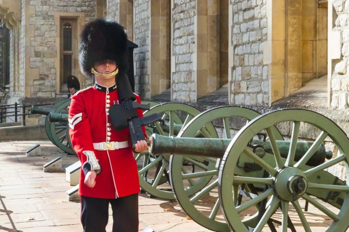 Guard in red uniform with black hat marches by cannons at a historic stone building.