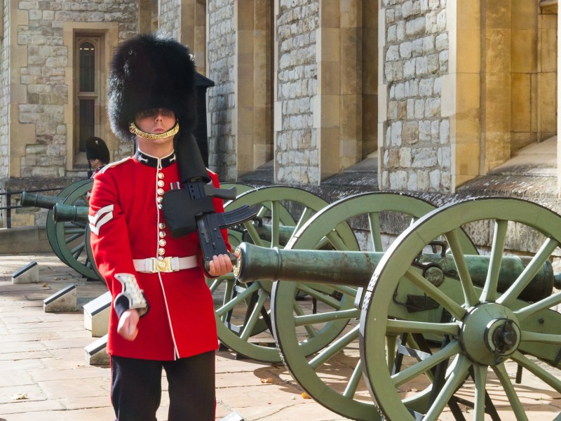 Guard in red uniform with black hat marches by cannons at a historic stone building.